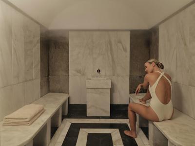 Frau in weißem Badeanzug sitzt in hellem Dampfbad mit Marmorbänken und gefalteten Handtüchern.