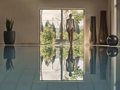 Frau steht im Badeanzug an einem großen Fenster mit Blick auf einen Garten, Poolspiegel glänzt.