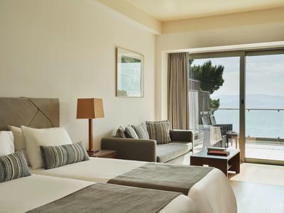 Helles Hotelzimmer mit zwei Betten, Sofa, Tisch und Meerblick durch große Fenster.