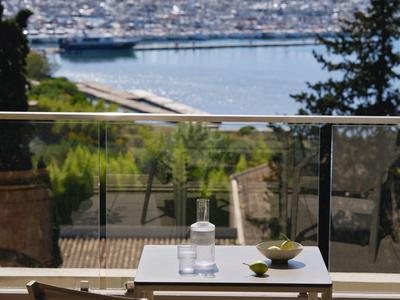 Balkon mit Tisch und zwei Stühlen, Blick auf Hafen mit Booten und bewaldete Hügel im Hintergrund.