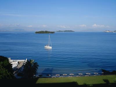 Blaues Meer mit Segelboot, Insel im Hintergrund und Strand mit Sonnenschirmen in einem Hotelresort.