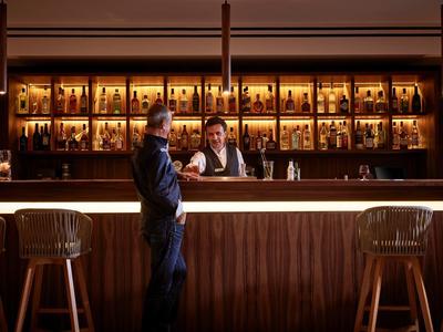 Eine Bar mit Holztheke, beleuchteten Regalen voller Flaschen und zwei Personen, die sich unterhalten.