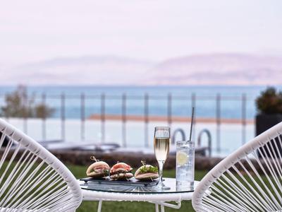 Zwei weiße Stühle an Tisch mit Hamburgern und Getränken, Meer und Berge im Hintergrund.
