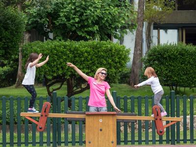 Drei Kinder spielen fröhlich auf einer großen, hölzernen Wippe im grünen Garten.
