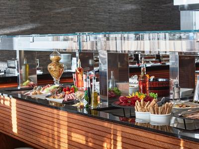 Buffet mit verschiedenen Speisen, frischem Gemüse und Brotkörben auf Holz-Theke unter Glasabdeckung.