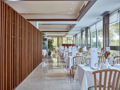 Modernes Restaurant mit langen weißen Tischen, Holzmöbeln und großen Glasfenstern, viel Tageslicht.