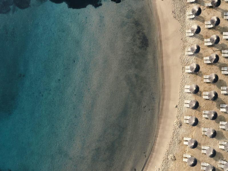 Luftaufnahme eines Strandes mit klarem blauem Wasser und Reihen von Sonnenschirmen auf hellem Sand.