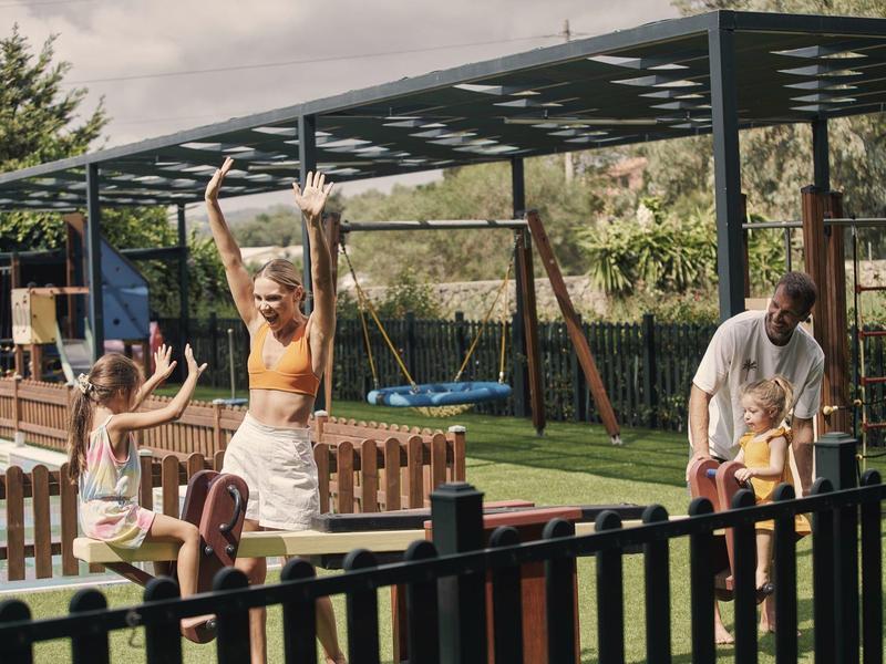Kinder spielen auf einer Holzwippe, begleitet von zwei Erwachsenen in einem Garten mit Spielplatz.