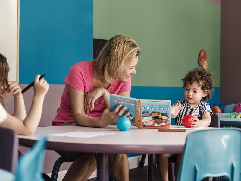 Frau und Kind sitzen an rundem weißen Tisch mit Spielzeug, bunter Wand im Hintergrund, Kinderzimmer.