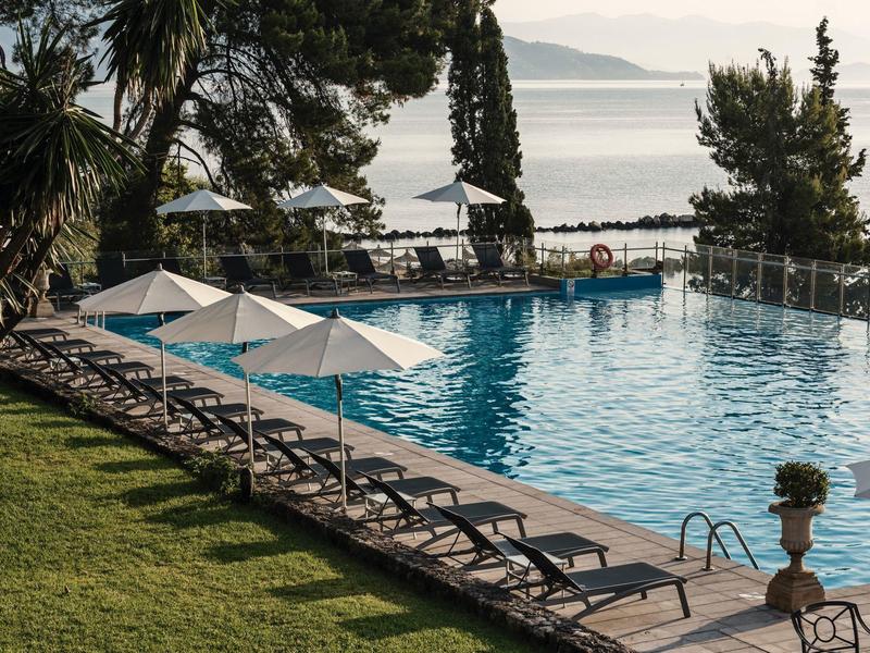 Pool mit Liegestühlen und Sonnenschirmen, umgeben von Bäumen, Blick auf ruhiges Meer im Hintergrund.