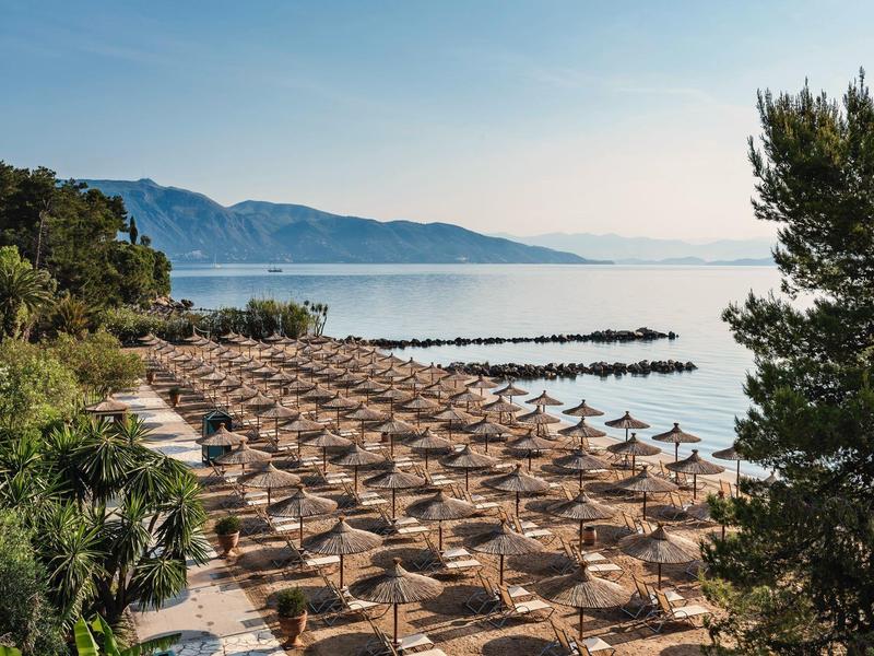 Leerer Strand mit Reihen aus Strohmuschel-Sonnenschirmen, ruhiges Meer und bewaldete Berge im Hintergrund.