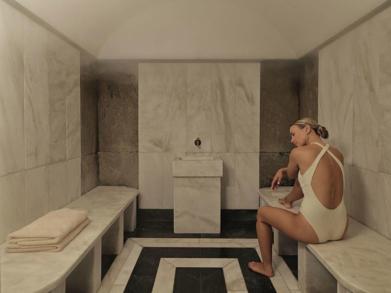 Frau in weißem Badeanzug sitzt in hellem Dampfbad mit Marmorbänken und gefalteten Handtüchern.