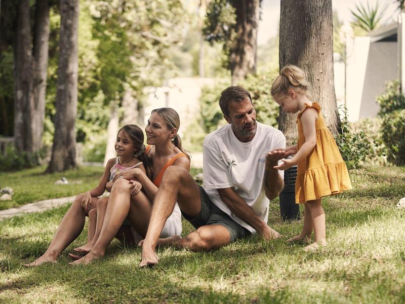 Familie entspannt im Park, sitzend auf Gras unter Bäumen an einem sonnigen Tag.