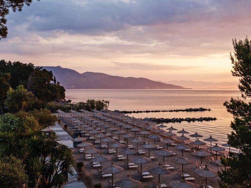 Strand mit Sonnenschirmen bei Sonnenaufgang, ruhiges Meer und Berge im Hintergrund.