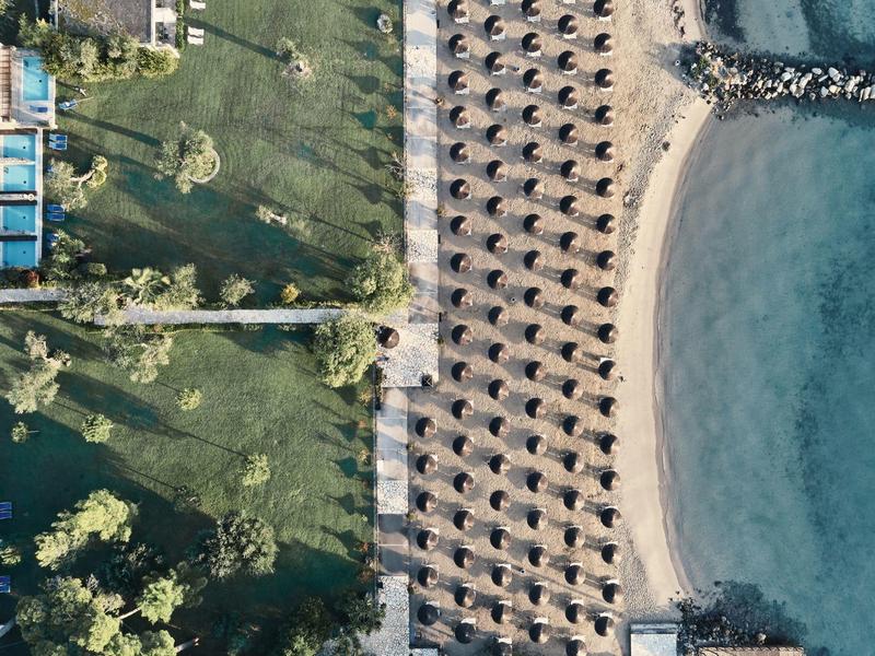 Luftaufnahme eines Strandes mit Reihen von Sonnenschirmen neben einem grünen Park und blauem Meer.