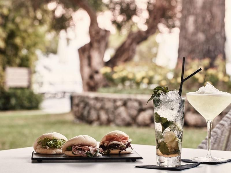 Drei Mini-Burger und zwei Drinks, ein Mojito und ein Cocktail, auf einem weißen Tisch im Freien.