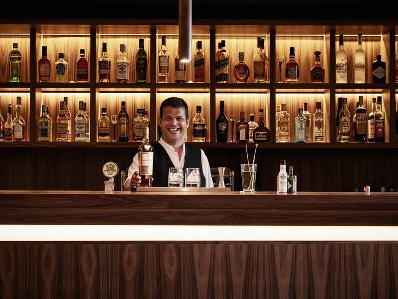 Ein Barkeeper steht hinter einer holzverkleideten Bar, umgeben von beleuchteten Regalen mit Flaschen.