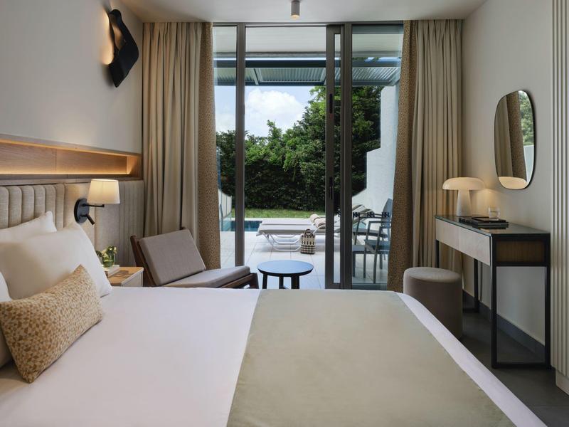 Modernes Hotelzimmer mit großem Bett, Sessel und Blick auf eine Terrasse mit Garten.