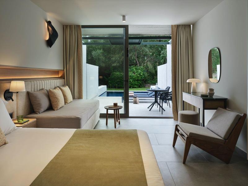 Helles Hotelzimmer mit Bett, Sofa, Stuhl und Blick auf Terrasse mit Garten und Pool.