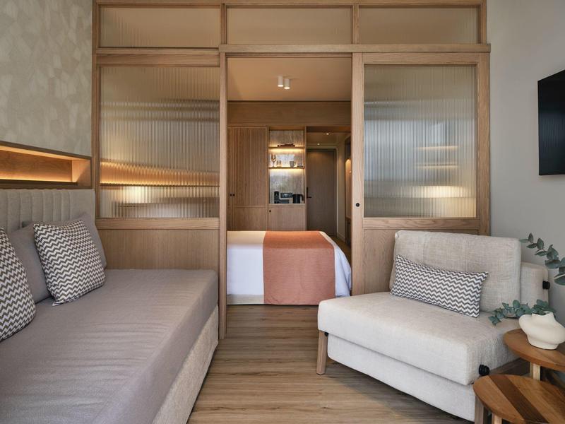 Modern eingerichtetes Hotelzimmer mit Sofa, Sessel und Blick auf das Schlafzimmer.