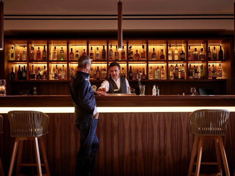 Eine Bar mit Holztheke, beleuchteten Regalen voller Flaschen und zwei Personen, die sich unterhalten.