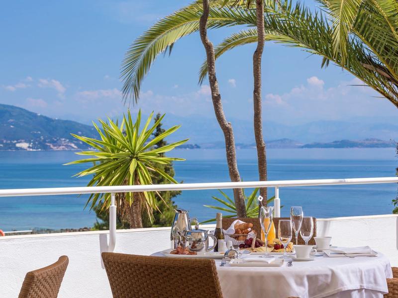 Blick auf gedeckten Tisch auf Terrasse mit Meer, Palmen und Bergen im Hintergrund.