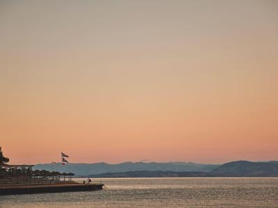 Sonnenuntergang über ruhigem Meer mit Strand und Bergen im Hintergrund.