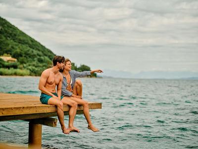 Zwei Männer sitzen am Holzsteg, blicken aufs Meer mit bewölktem Himmel und grünen Hügeln.