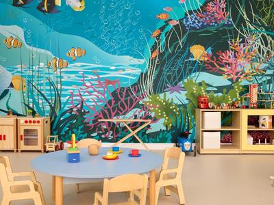 Kindergartenraum mit rundem blauen Tisch, Holzstühlen, Regal und bunter Unterwassermalerei an der Wand.