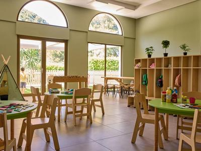 Heller, grüner Gruppenraum mit Holzstühlen, Tischen und Spielzeug-Regal für Kinder.