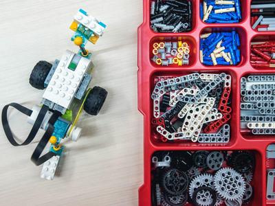 Lego-Rennwagen aus weißen, schwarzen und grünen Steinen neben rot-schwarzer Sortierbox mit Zahnrädern und Stiften.