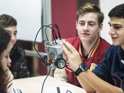 Vier Jugendliche betrachten gespannt einen kleinen grauen Roboter mit Rädern an einem Tisch.