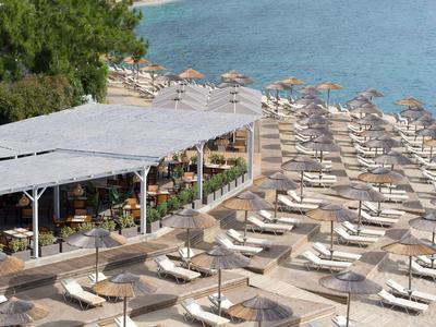 Strand mit vielen Liegestühlen und Sonnenschirmen neben einem überdachten Sitzbereich am blauen Wasser.