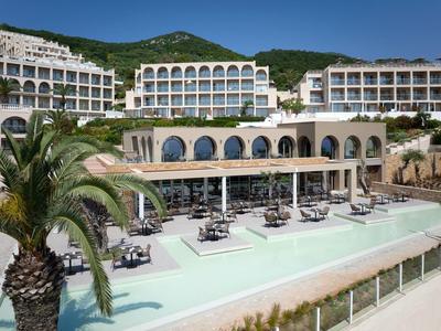 Großes Hotel mit mehreren weißen Gebäuden, Pool mit Sonnenschirmen und Palmen davor, grüner Berg im Hintergrund.