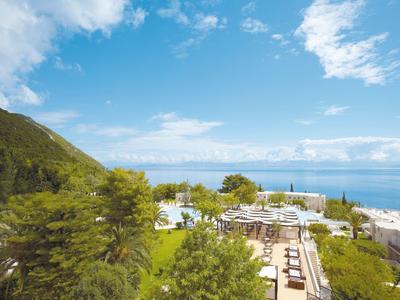 Blick auf einen grünen Hügel, Swimmingpools und weiße Sonnenschirme am Meer unter blauem Himmel.