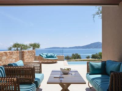 Terrasse mit blauen Polstersofas, Holzmöbeln und Blick auf das Meer und Berge bei klarem Himmel.