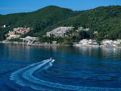 Ein Boot fährt auf dem blauen Wasser vor bewaldeten Hügeln und einer Küstenstadt.