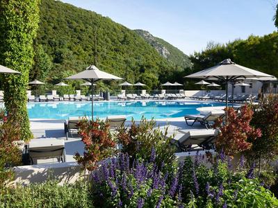 Großer Pool mit Liegestühlen, Sonnenschirmen und grünen Bergen im Hintergrund bei klarem Himmel.