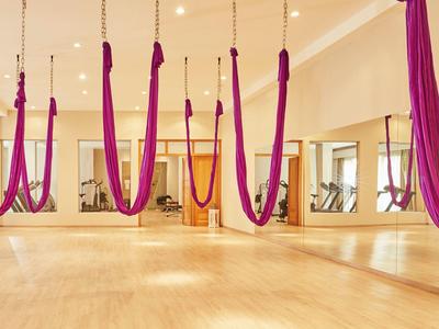 Sala de yoga luminosa con suelo de madera, grandes espejos y telas colgantes de yoga aéreo en morado y naranja.