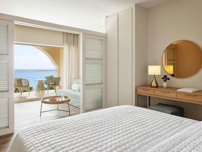 Helles Schlafzimmer mit weißer Bettdecke, Holzmöbeln und Balkon mit Meerblick bei Tageslicht.