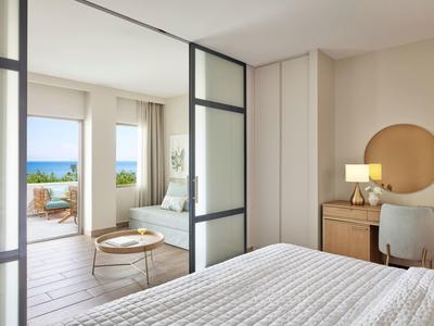 Helles Schlafzimmer mit weißer Bettdecke, Holzschreibtisch, Stuhl und Meerblick durch große Fenster.