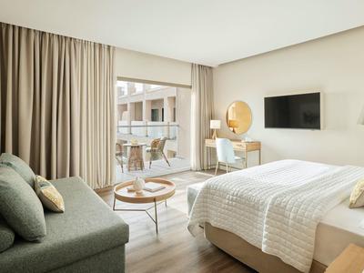 Helles, modernes Schlafzimmer mit großem Bett, grauem Sofa, hellem Holztisch und Balkon mit Stühlen.