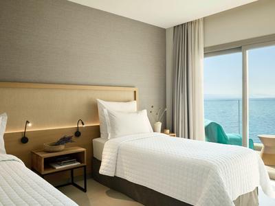 Helles Hotelzimmer mit zwei Einzelbetten, Nachttisch, Leselampen und Meerblick durch großes Fenster.