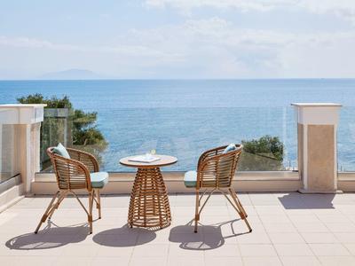 Balkon mit Glasgeländer, zwei Rattanstühlen und Tisch mit Meerblick unter blauem Himmel.