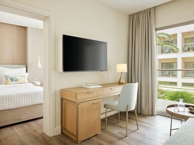 Moderner Raum mit Schreibtisch, Stuhl, großem Fernseher, Bett im Nebenraum und großem Fenster.