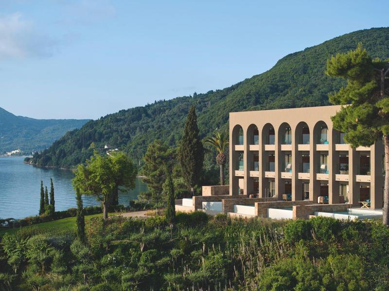 Modernes Gebäude mit Bögen neben bewaldetem Hügel und ruhigem See unter blauem Himmel.