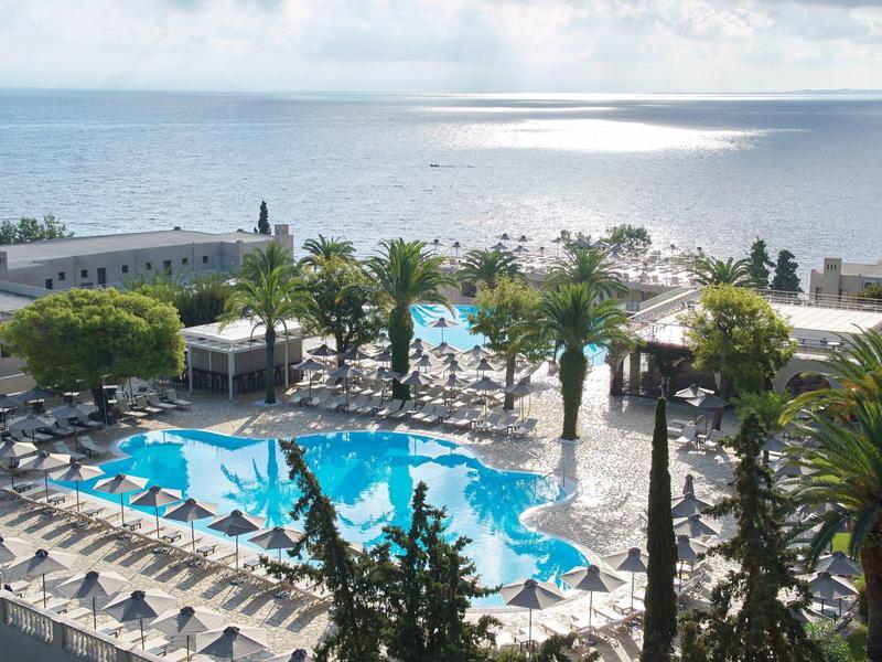 Großer Pool mit Liegestühlen und Palmen neben einem hellen Meer unter bewölktem Himmel.