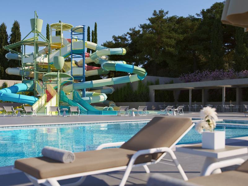 Schwimmbad mit Wasserrutschen, Liegestühlen und klar blauem Himmel im Hintergrund.