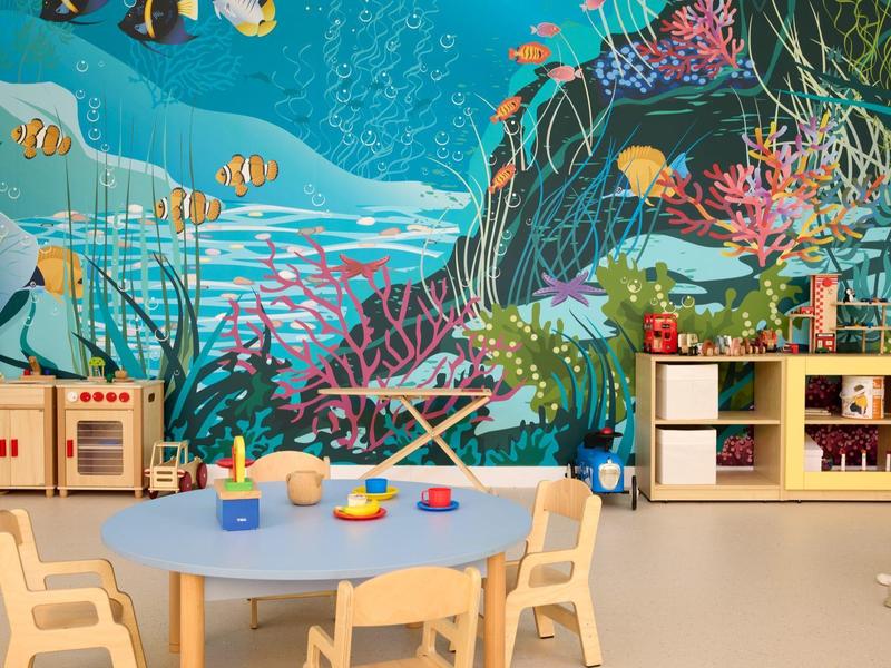 Kindergartenraum mit rundem blauen Tisch, Holzstühlen, Regal und bunter Unterwassermalerei an der Wand.