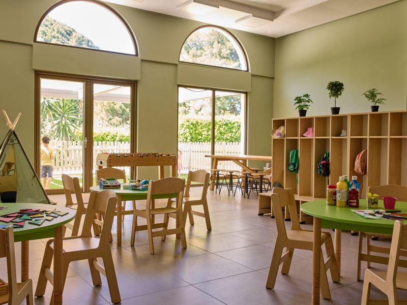 Heller, grüner Gruppenraum mit Holzstühlen, Tischen und Spielzeug-Regal für Kinder.